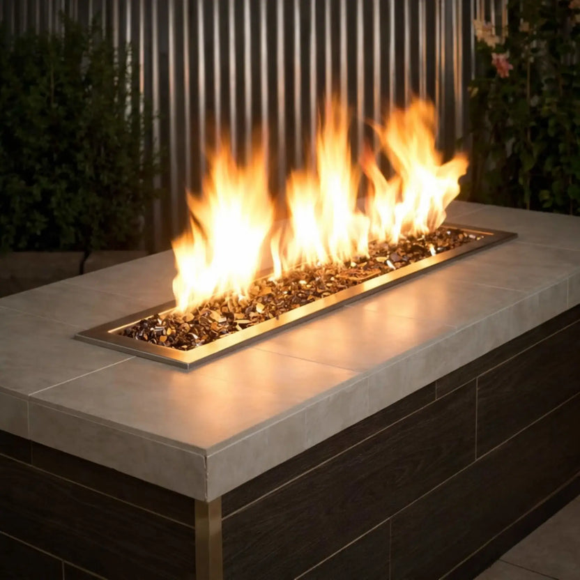 Modern linear outdoor fire pit burner filled with American Fire Products 1/2 inch premium reflective bronze fire glass, producing tall golden flames across a sleek stainless steel insert set into a contemporary tile-top patio table.