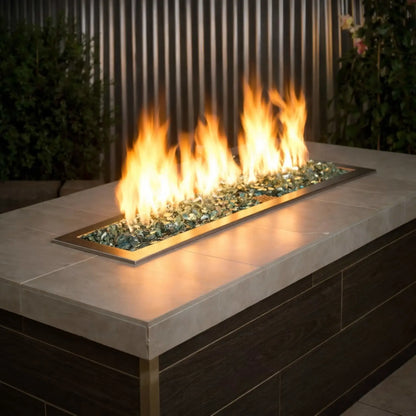 Modern linear outdoor gas fire pit filled with American Fire Products 1/2 inch premium reflective evergreen green fire glass, producing tall golden flames across a sleek stainless steel burner insert set into a contemporary patio table.