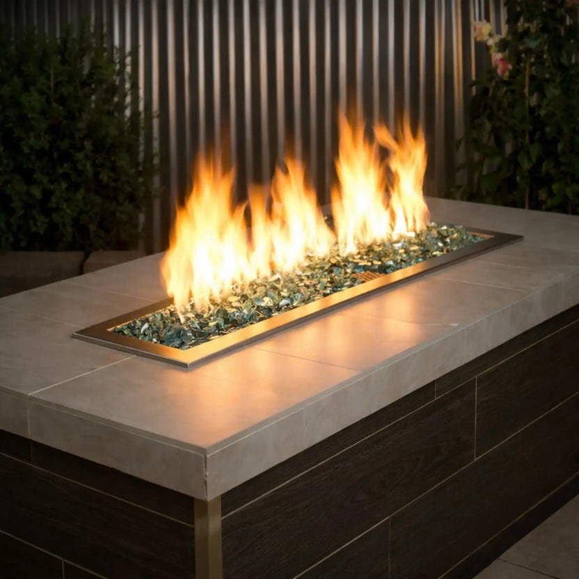 Modern linear outdoor gas fire pit filled with American Fire Products 1/2 inch premium reflective evergreen green fire glass, producing tall golden flames across a sleek stainless steel burner insert set into a contemporary patio table.