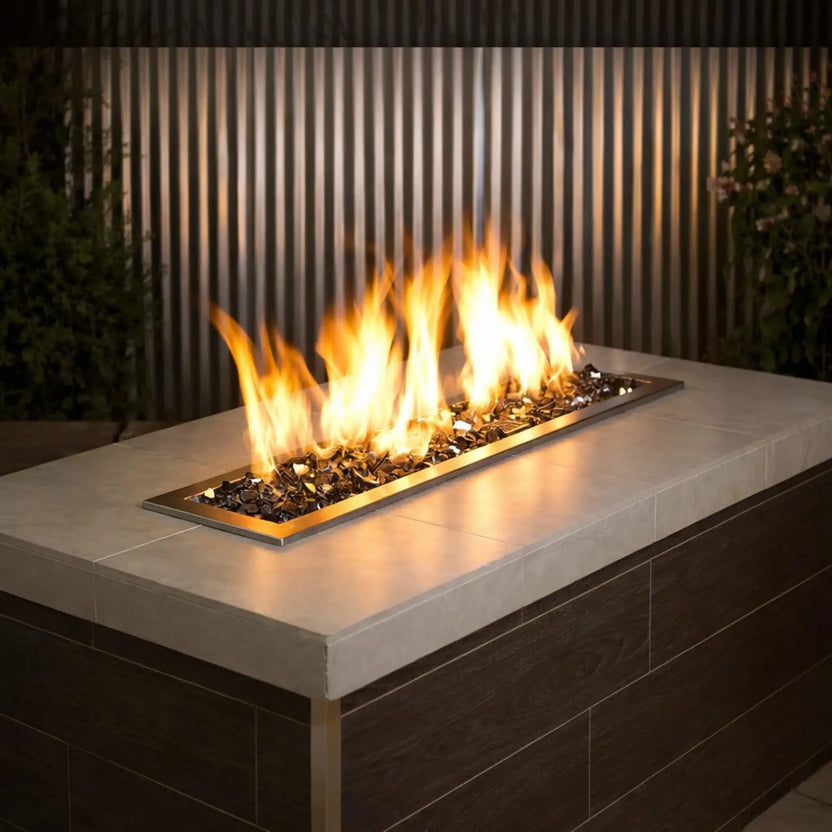 Modern linear outdoor gas fire pit filled with 1/2 inch premium reflective gray fire glass by American Fire Products, featuring tall golden flames rising from a stainless steel burner set into a contemporary stone-topped fire table with a sleek wood-paneled base.
