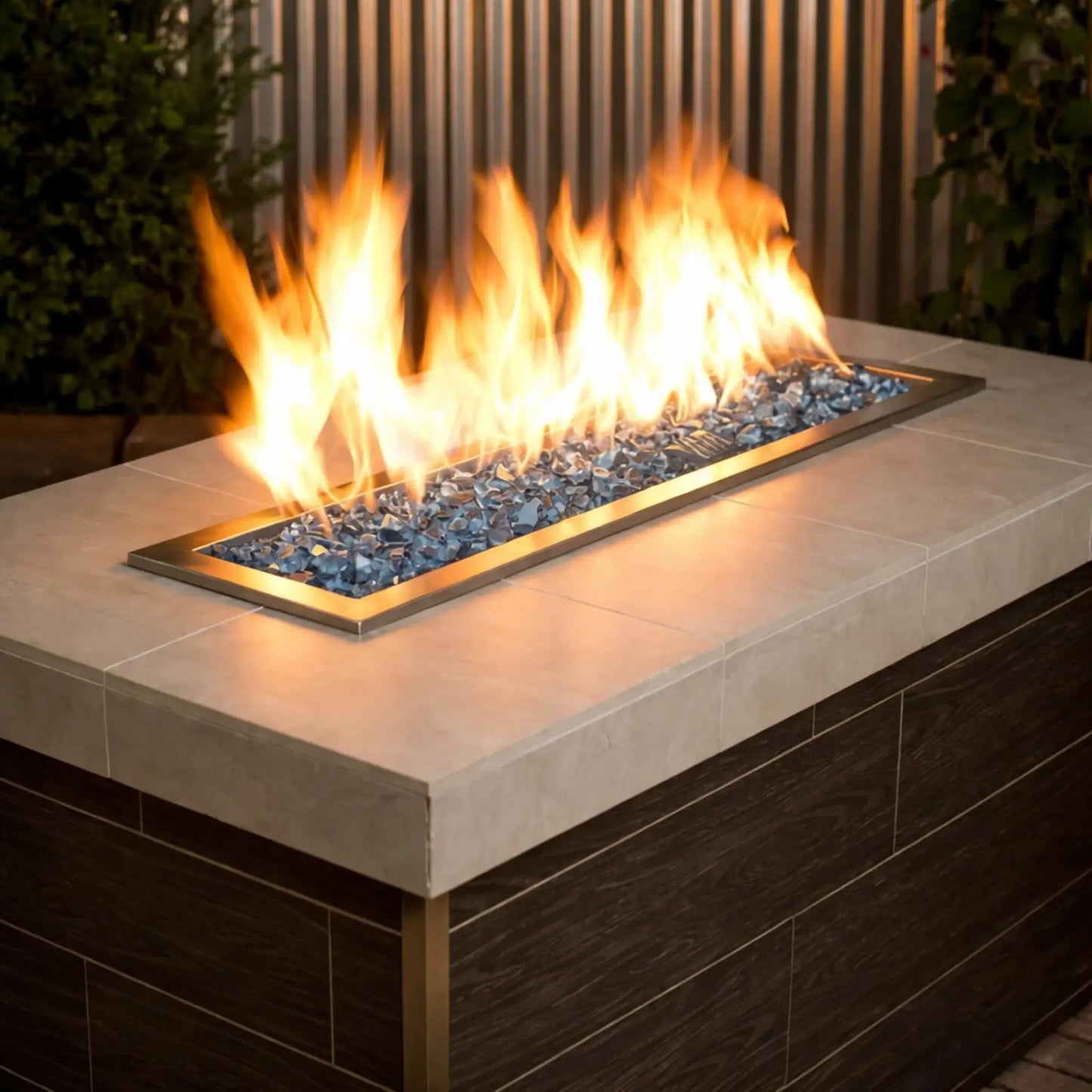 Contemporary rectangular gas fire pit filled with American Fire Products 1/2 inch premium reflective Pacific blue fire glass, where bright yellow flames dance across a stainless steel burner pan set into a modern patio fire table.