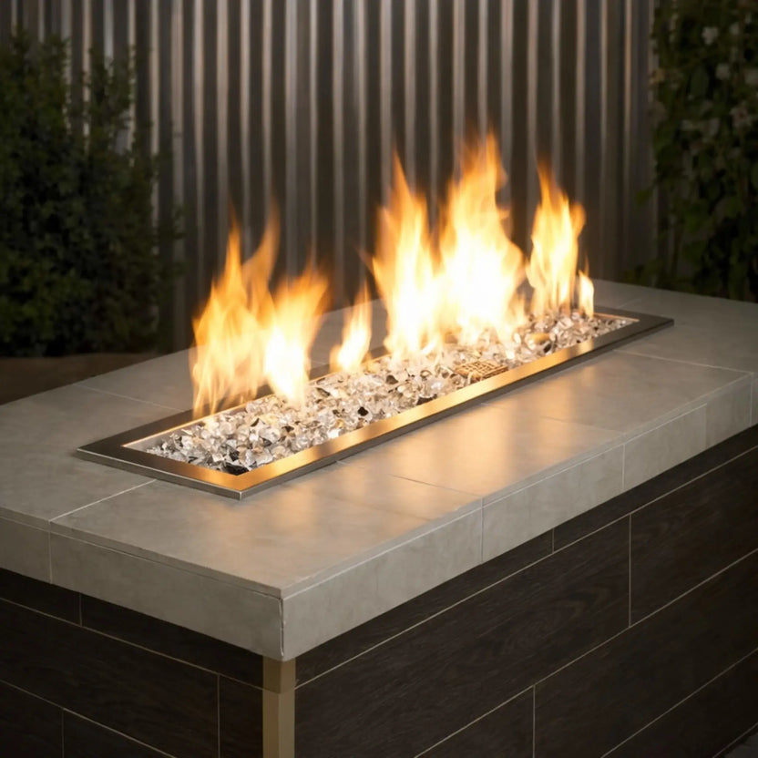 Sleek linear outdoor gas fire pit filled with American Fire Products 1/2 inch premium reflective Starfire ultra clear titanium fire glass, producing tall golden flames across a stainless steel burner insert set into a contemporary stone fire table.