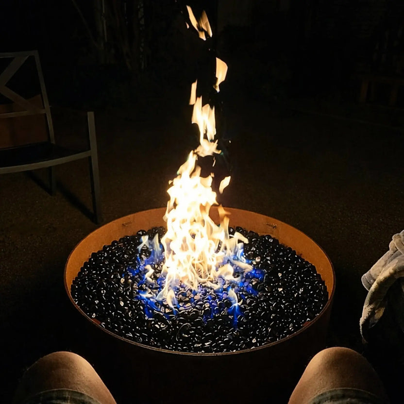 Nighttime patio fire bowl with black tempered glass beads glowing beneath tall yellow flames and blue fire at the base, creating a dramatic outdoor ambiance.