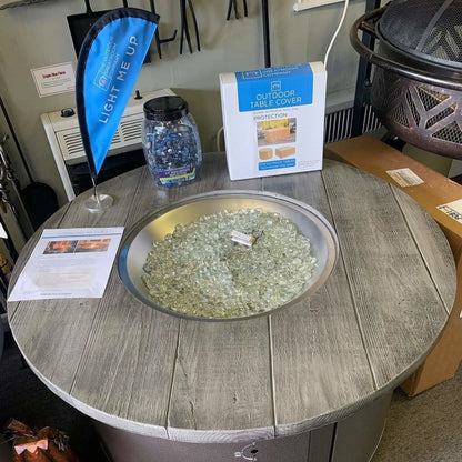 Round outdoor fire pit table display in a showroom setting, filled with clear tempered glass luster beads inside a brushed metal burner ring, surrounded by product signage and accessories.