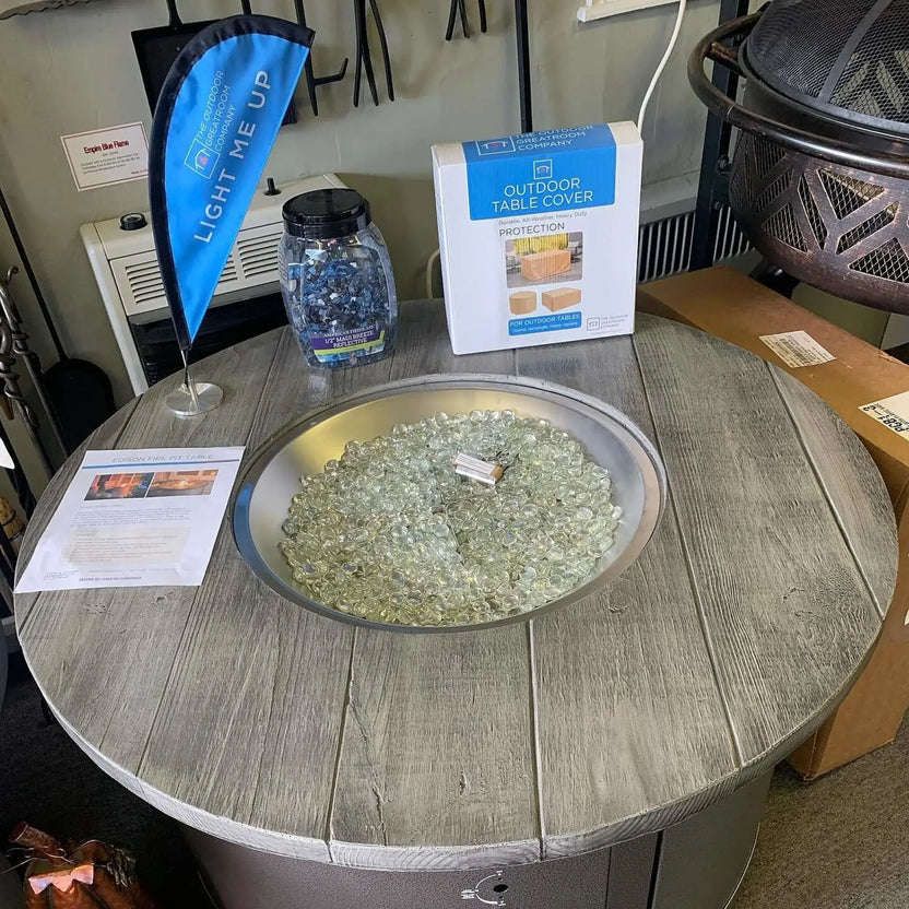 Round outdoor fire pit table display in a showroom setting, filled with clear tempered glass luster beads inside a brushed metal burner ring, surrounded by product signage and accessories.