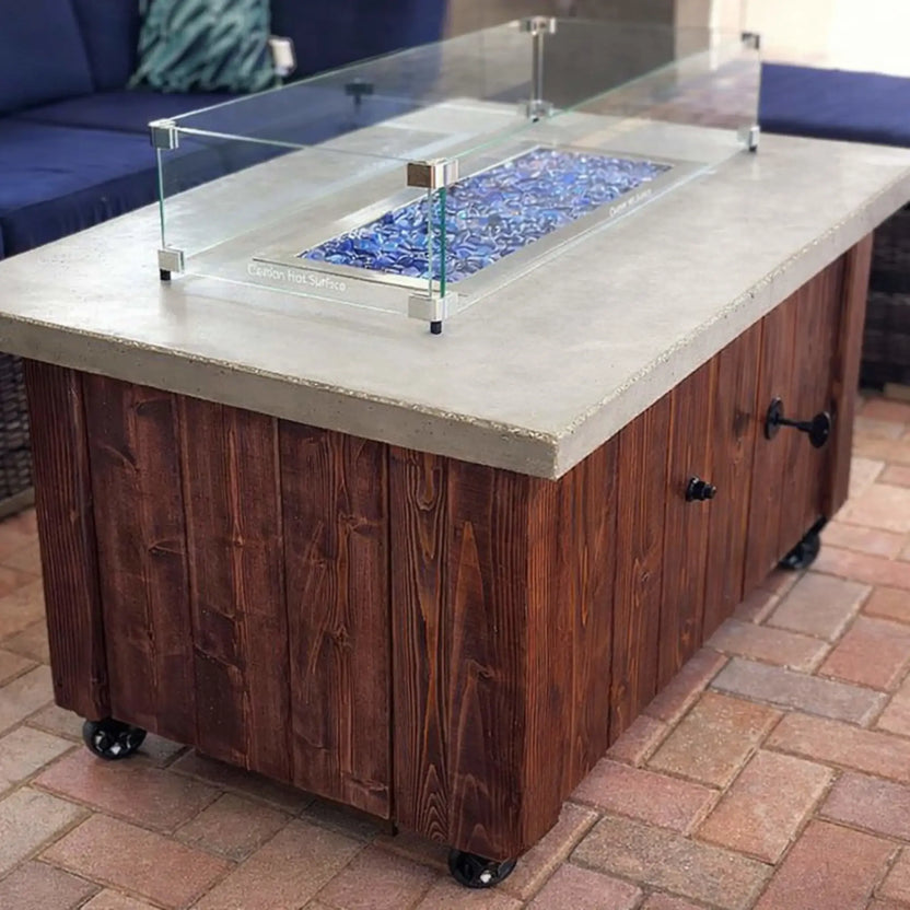 Rectangular patio fire table with a concrete top and rich wood base on casters, featuring a linear burner filled with blue tempered glass beads protected by a clear glass wind guard.