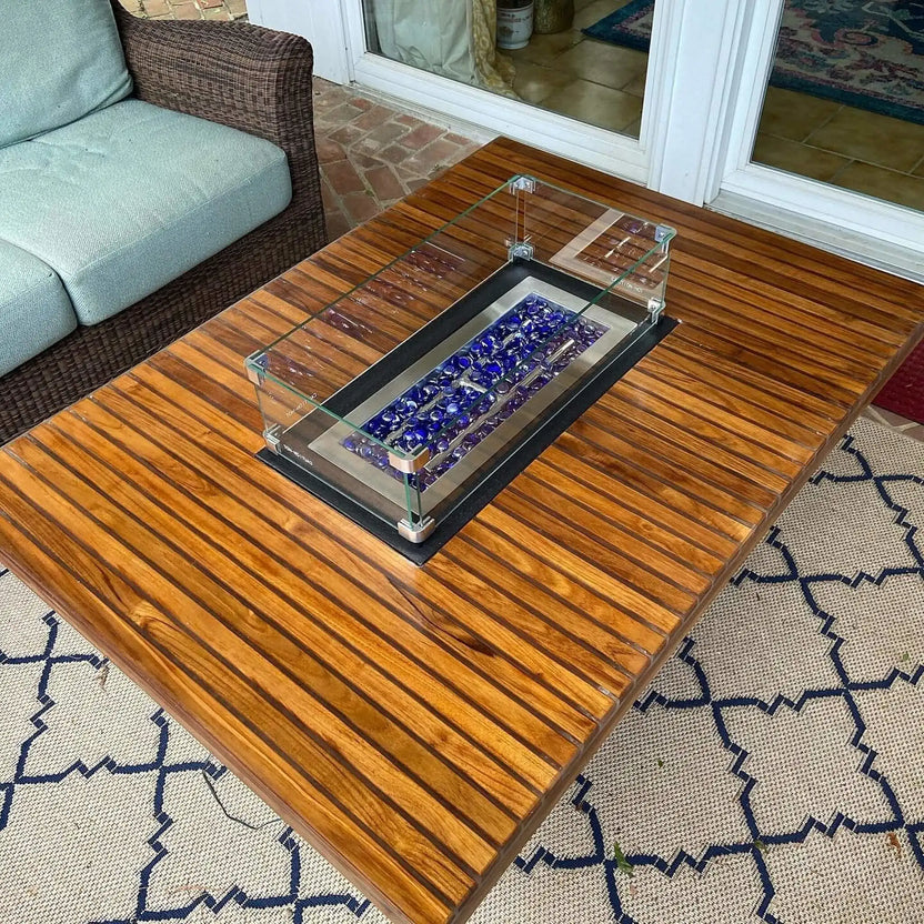 Slatted wood outdoor coffee table with a stainless steel linear burner filled with royal blue glass beads, topped with a clear glass wind guard on a covered patio.