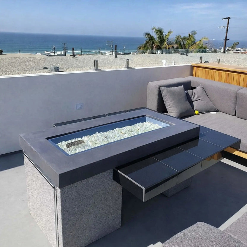 Modern rooftop patio overlooking the ocean with a sleek rectangular fire table filled with clear fire glass beads, set beside cushioned outdoor seating and black tile accents.