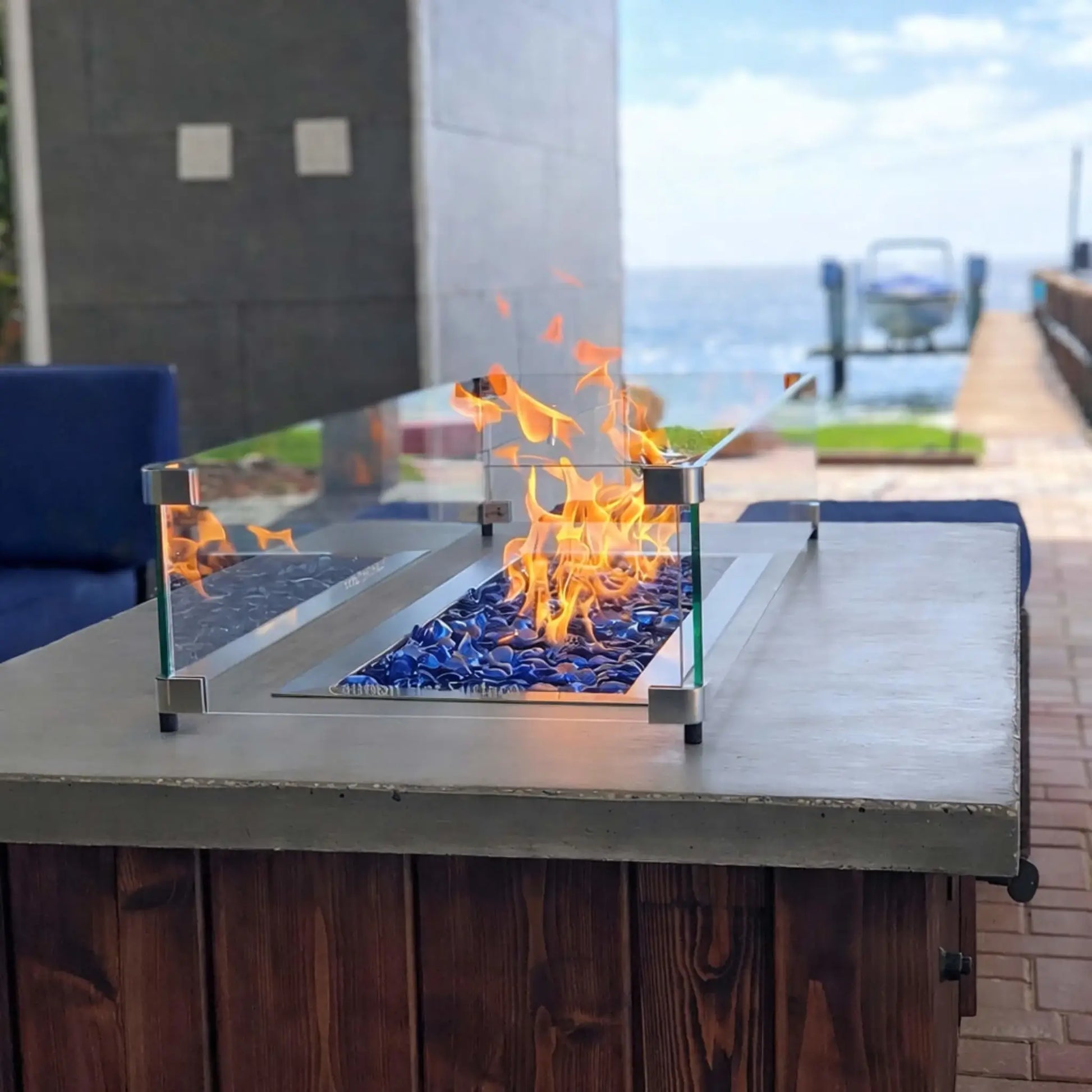 Contemporary linear fire feature built into a stone bench, glowing with bright flames over deep blue tempered glass beads in a landscaped backyard with tiered rock retaining walls.