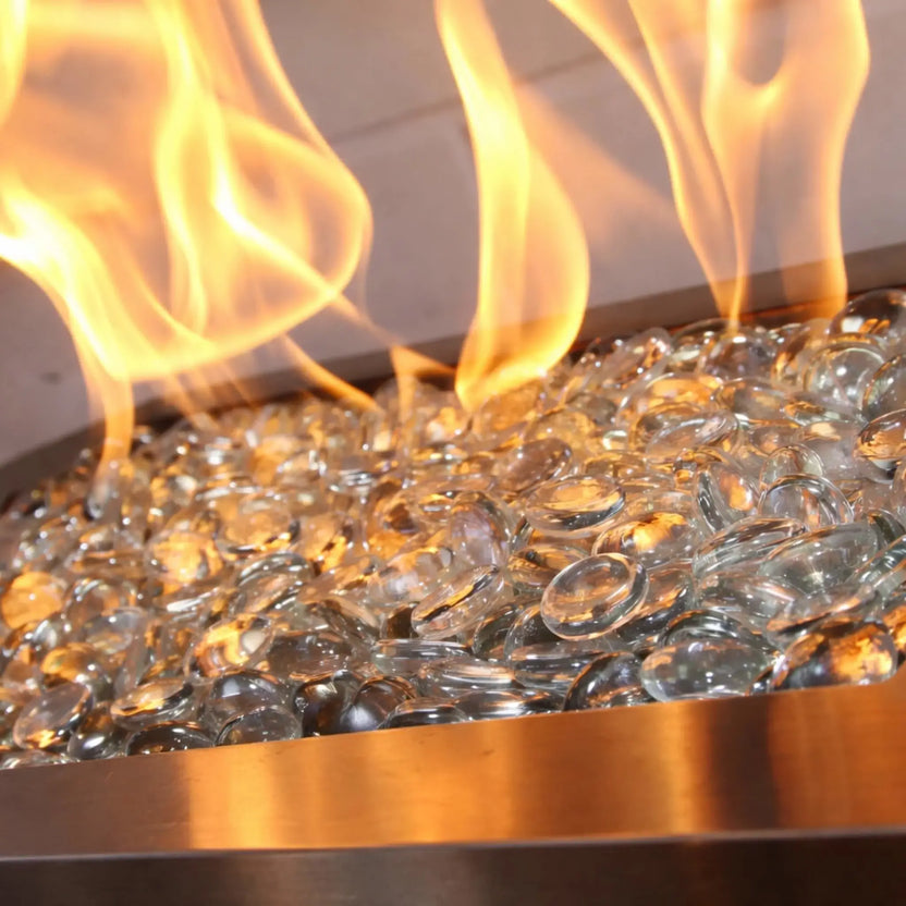 Tight detail of golden flames rising from clear fire glass beads in a brushed metal burner tray, highlighting the polished finish and light-catching texture of the tempered glass media.