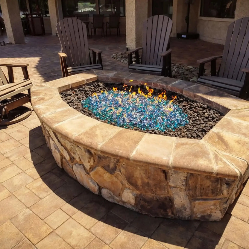 Curved stone outdoor gas fire pit filled with shimmering aqua blue tempered glass luster beads, surrounded by dark lava rock and Adirondack chairs on a paved backyard patio.