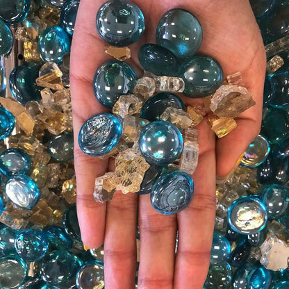 Hand holding a mix of aqua blue luster fire glass beads and clear crushed fire glass, showing the smooth rounded drops and reflective tempered finish up close.