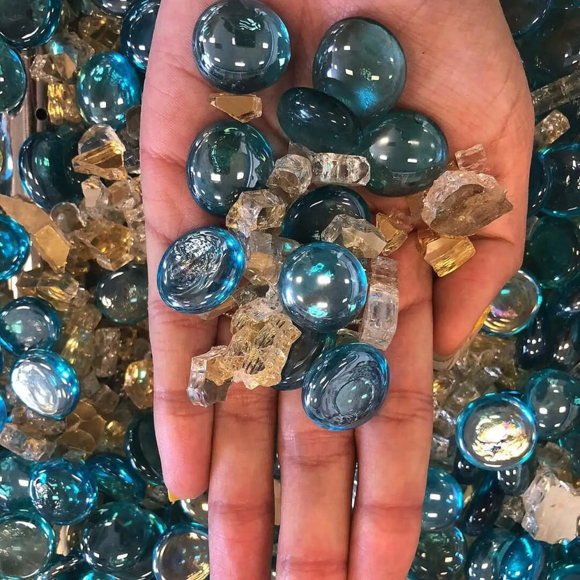Hand holding a mix of aqua blue luster fire glass beads and clear crushed fire glass, showing the smooth rounded drops and reflective tempered finish up close.