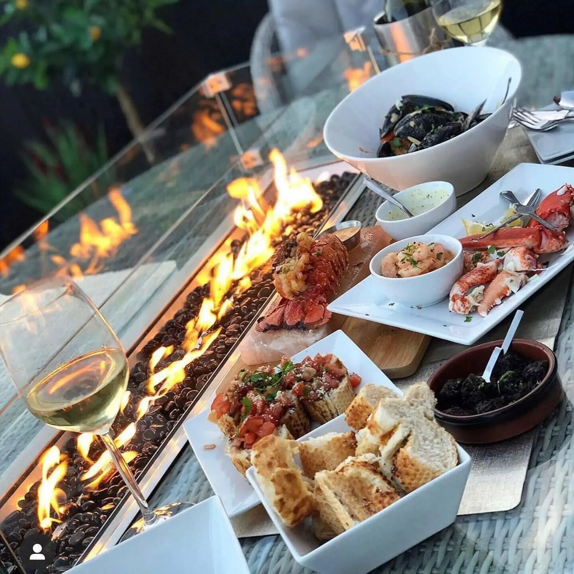 Outdoor dining table with a linear gas fire feature filled with glossy black glass beads, set beside plated seafood, bread, and wine for an evening patio setting.
