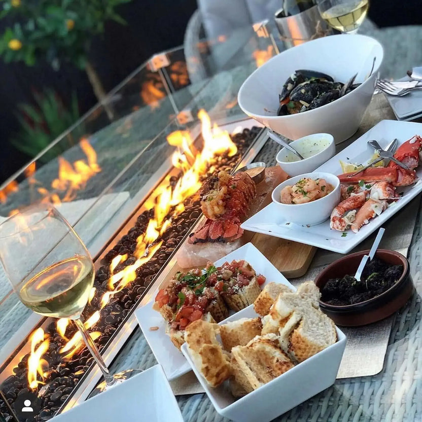 Outdoor dining table with a linear gas fire feature filled with glossy black glass beads, set beside plated seafood, bread, and wine for an evening patio setting.