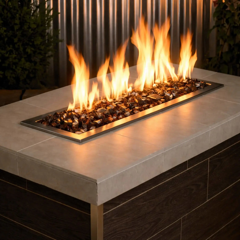 Linear stainless steel gas fire burner filled with Auburn tempered fire glass, producing tall golden flames across a modern outdoor fire table with a smooth concrete top and dark wood base.