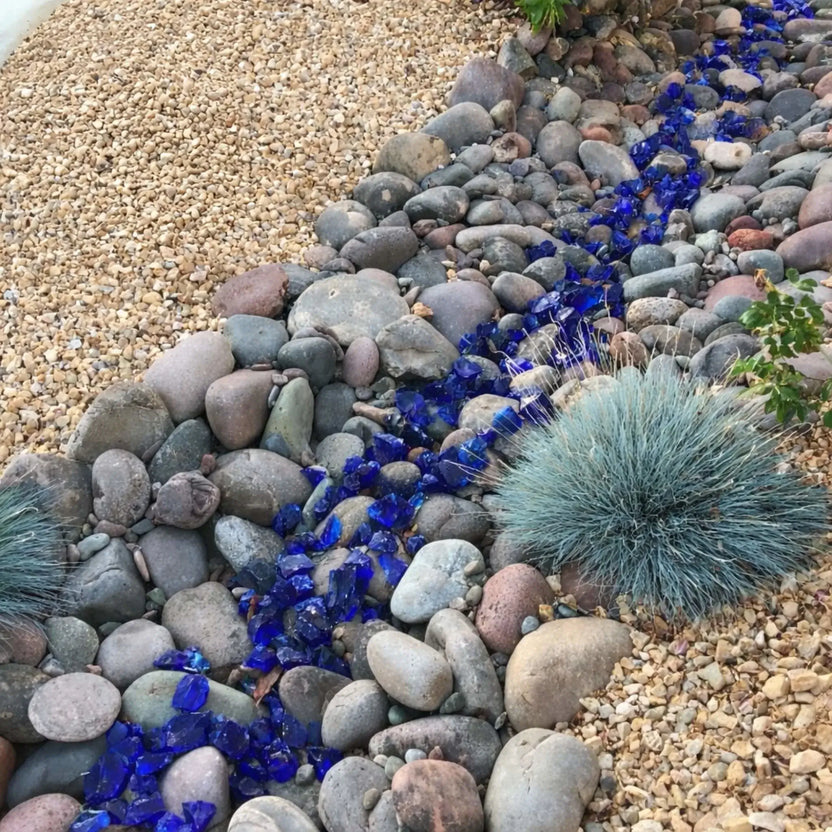 Decorative landscaping installation using dark blue fire glass as a river-style accent between natural stones and gravel, adding bold color contrast to a xeriscape garden design.