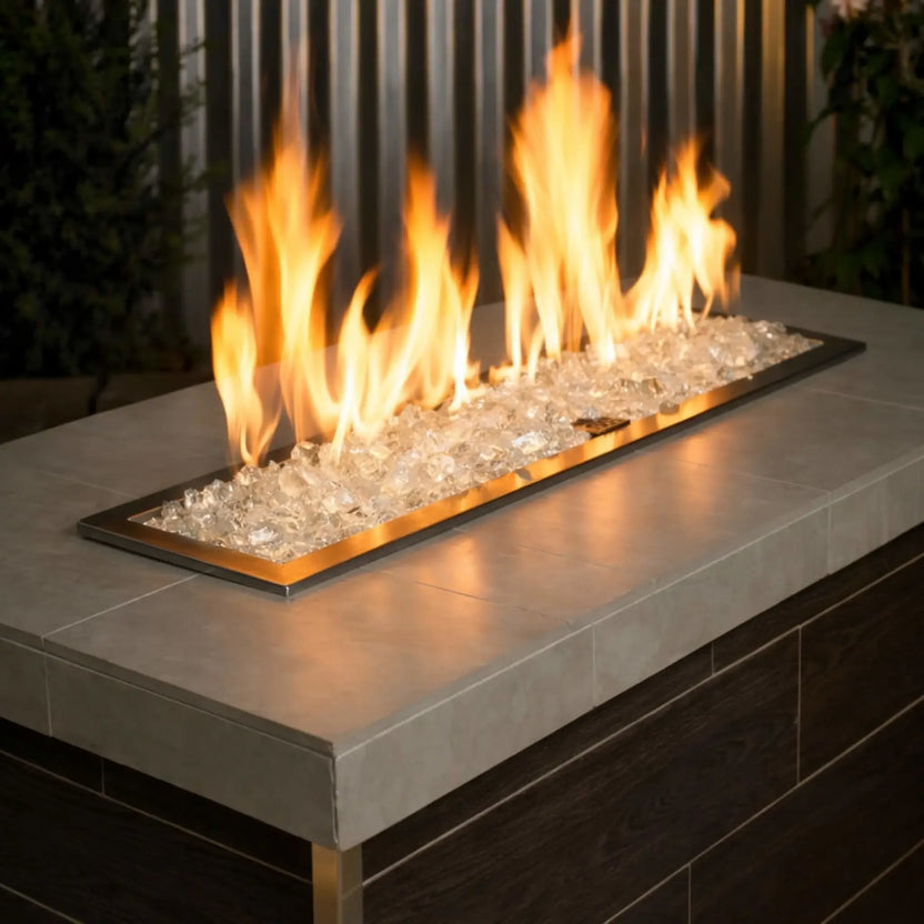 Linear stainless steel gas fire burner filled with Ice White tempered fire glass, producing warm golden flames across a sleek outdoor fire table with a concrete top and dark wood base.