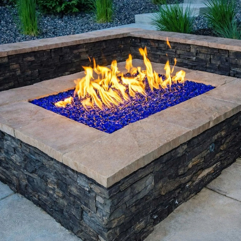 Square stone fire pit featuring light blue fire glass beneath lively flames, surrounded by stacked stone walls and landscaped greenery in a backyard patio setting.