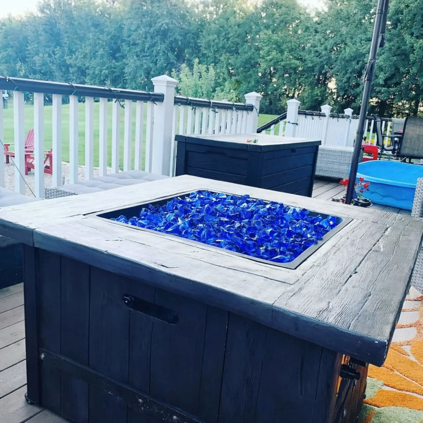 Outdoor deck fire table filled with light blue tempered fire glass, set against white railing and backyard lawn, creating a vibrant centerpiece for a residential patio space.