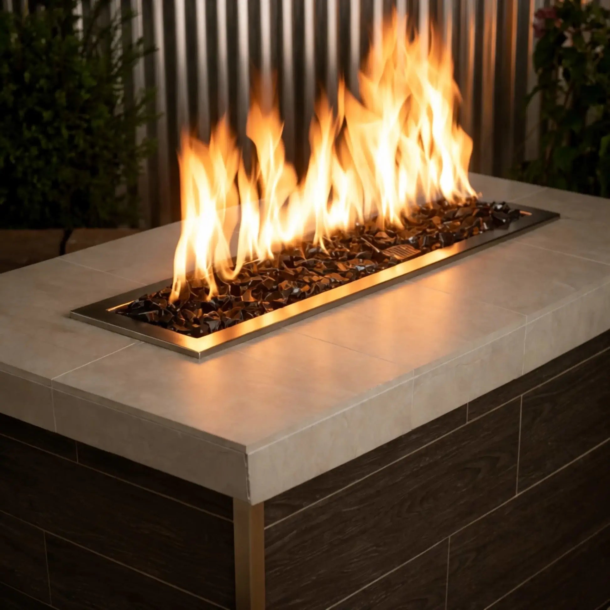 Modern linear gas fire table filled with Onyx Black fire glass, with tall golden flames rising from a stainless steel burner set into a sleek concrete tabletop.