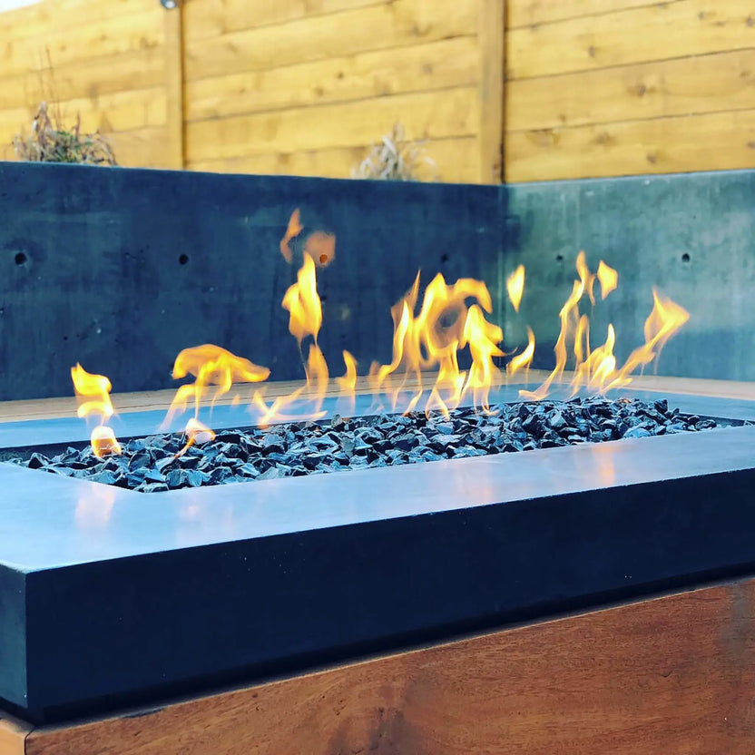 Contemporary outdoor fire feature with black tempered fire glass burning beneath bright flames, framed by a minimalist concrete surround and wood-paneled base.