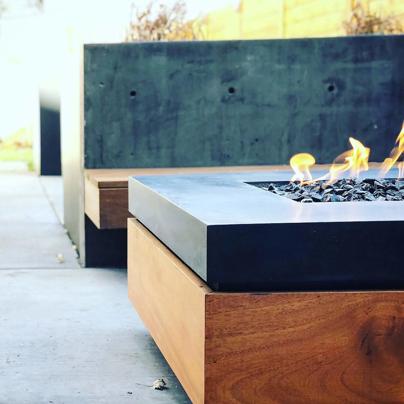 Rectangular fire pit with Onyx Black fire glass set into a dark metal surround, flames dancing along the surface in a modern patio setting with wood fencing backdrop.