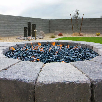 Round stone fire pit filled with black tempered fire glass, small orange flames flickering across the surface in a landscaped backyard with block walls and decorative rock accents.
