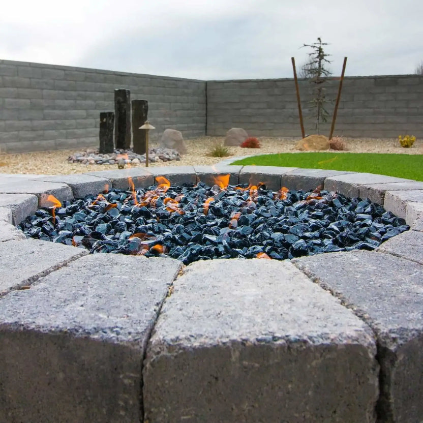 Round stone fire pit filled with black tempered fire glass, small orange flames flickering across the surface in a landscaped backyard with block walls and decorative rock accents.