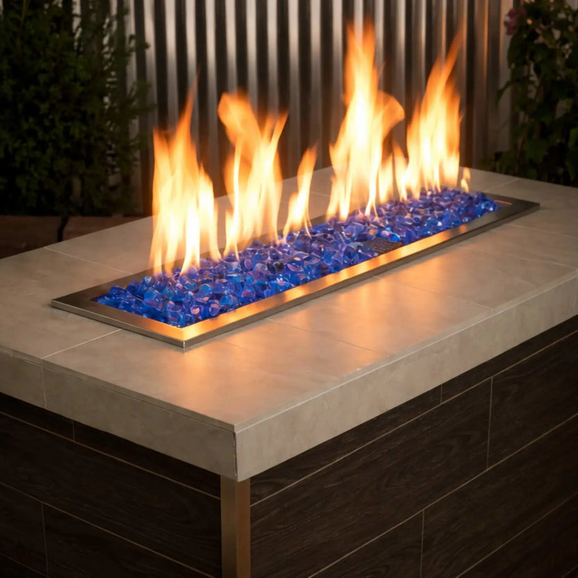 Modern linear gas fire table filled with turquoise blue tempered fire glass, with tall golden flames rising from a stainless steel burner set into a smooth concrete top and dark wood base.