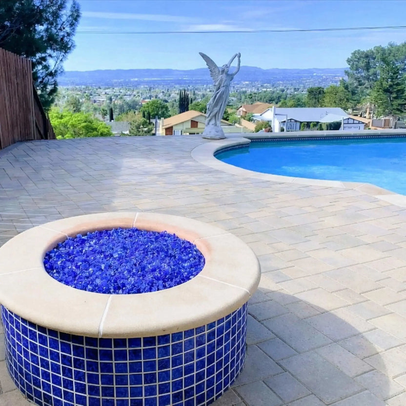 Round outdoor fire bowl packed with vibrant turquoise blue fire glass, set beside a curved swimming pool on a spacious paver patio overlooking a hillside neighborhood and valley views.