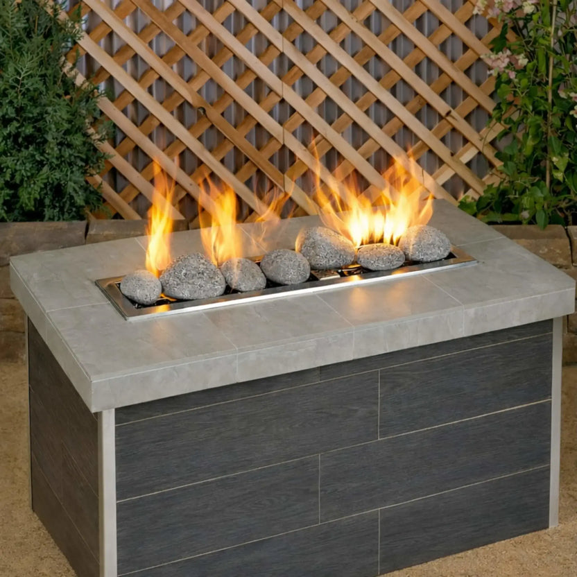 Rectangular outdoor gas fire table featuring extra large gray lava stones from American Fire Products arranged along a linear burner, with bright yellow flames set against a backyard lattice wall backdrop.