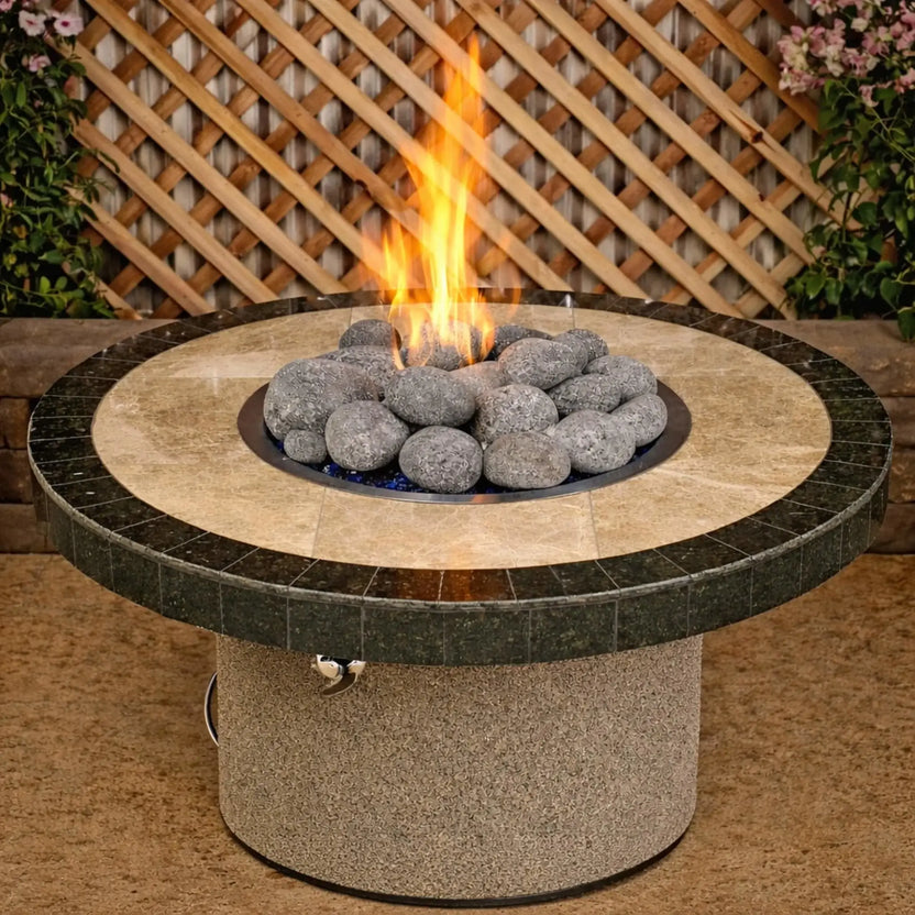 Round patio fire pit table filled with extra large American Fire Products lava stones surrounding a central gas flame, showcasing a natural stone top and backyard setting with wood lattice and greenery.