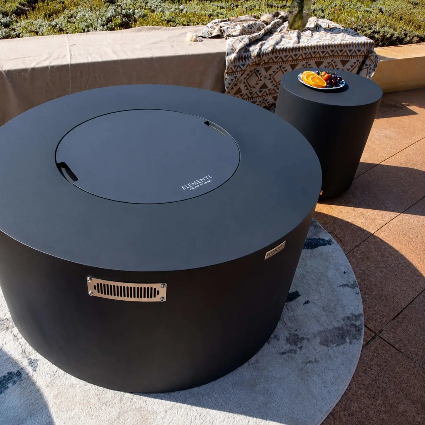Elementi Art of Living Pacaya Dark Grey round GFRC concrete fire pit table shown with its matching burner cover in place, creating a sleek outdoor coffee table appearance. The hidden propane fire pit features a smooth circular design, integrated ventilation panel, and concealed drawer system for propane tank storage. Positioned on a round outdoor rug beside a matching side table, this luxury patio centerpiece overlooks a landscaped terrace with modern outdoor décor.