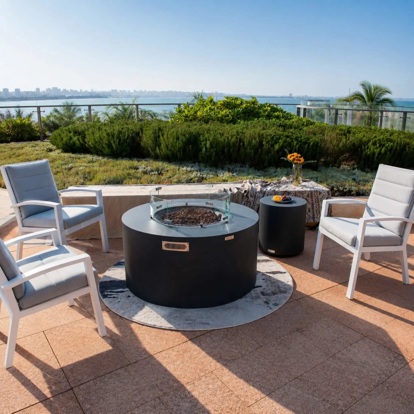 Full lifestyle scene featuring the Elementi Art of Living Pacaya Dark Grey round hidden propane fire pit table centered within a contemporary outdoor seating arrangement. The GFRC concrete fire table includes a glass wind guard, lava rock burner media, concealed propane drawer system, and matching side table, creating a sophisticated waterfront patio gathering space with panoramic landscape views.