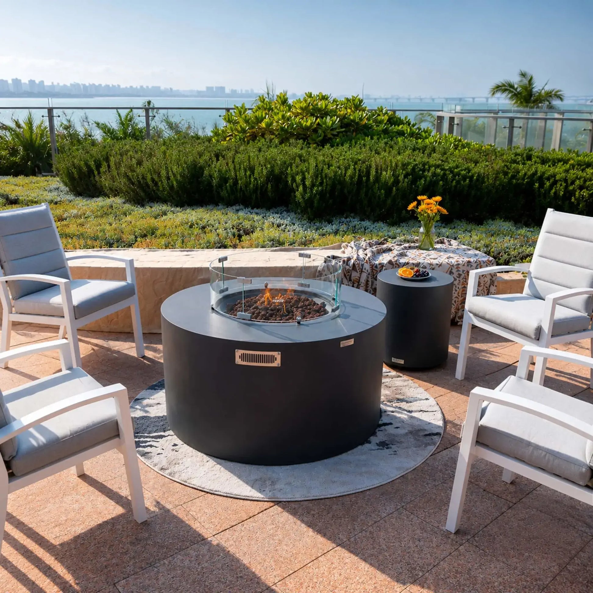 Elementi Art of Living Pacaya Dark Grey round GFRC concrete fire pit table showcased as the centerpiece of a luxury outdoor lounge. Featuring a hidden propane fire pit, circular glass wind guard, lava rock burner media, and concealed drawer system, the fire table is surrounded by four cushioned patio chairs on a round outdoor rug overlooking landscaped gardens and a waterfront view.