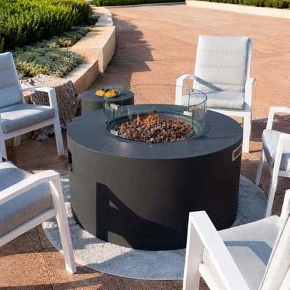 Angled view of the Elementi Art of Living Pacaya Dark Grey round fire pit table with active flames protected by a tempered glass wind guard. The GFRC concrete fire table sits within a modern patio seating arrangement featuring four cushioned armchairs, lava rock media, hidden propane storage, and a matching side table, creating a sophisticated outdoor gathering space.