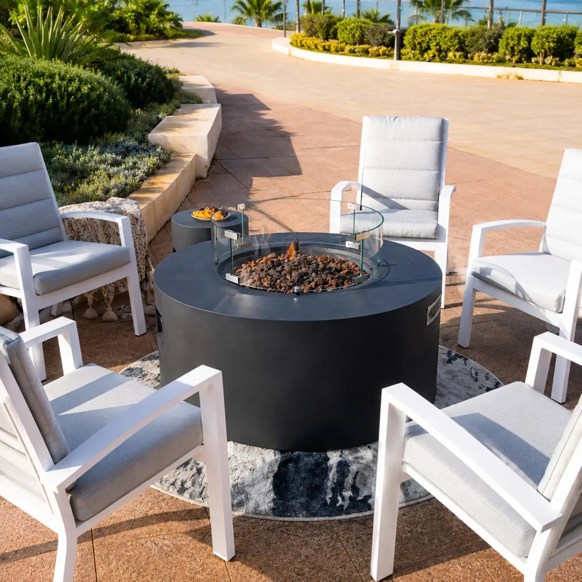 Elementi Art of Living Pacaya Dark Grey hidden propane fire pit table positioned at the center of a circular conversation area. The round GFRC concrete fire table features a glass wind guard surrounding a lava rock burner and is paired with four modern patio chairs, offering a comfortable and stylish outdoor entertainment setting beside lush landscaping and waterfront scenery.