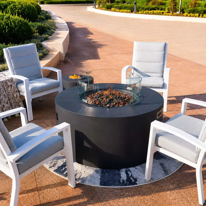 Wide lifestyle image of the Elementi Art of Living Pacaya Dark Grey round fire pit table with visible flames and a tempered glass wind guard. The hidden propane fire feature is arranged with modern white-framed outdoor chairs on a spacious patio overlooking water views, showcasing the table’s contemporary design, concealed fuel storage, and luxury outdoor living appeal.