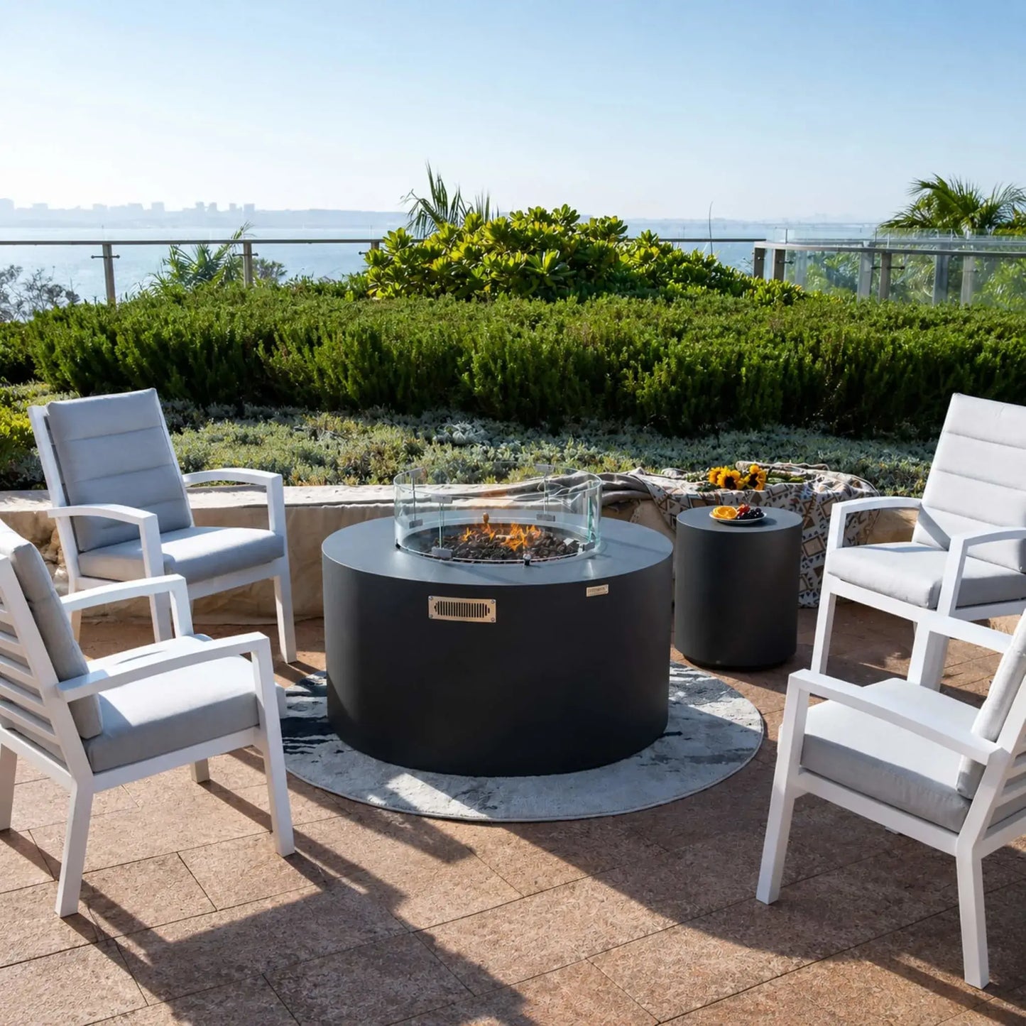 Front-facing view of the Elementi Art of Living Pacaya Dark Grey GFRC concrete fire pit table surrounded by four cushioned lounge chairs. The hidden propane fire pit features a circular glass wind guard, natural lava rock burner media, and integrated storage, while a matching side table with flowers and fruit accents enhances the upscale waterfront patio atmosphere.