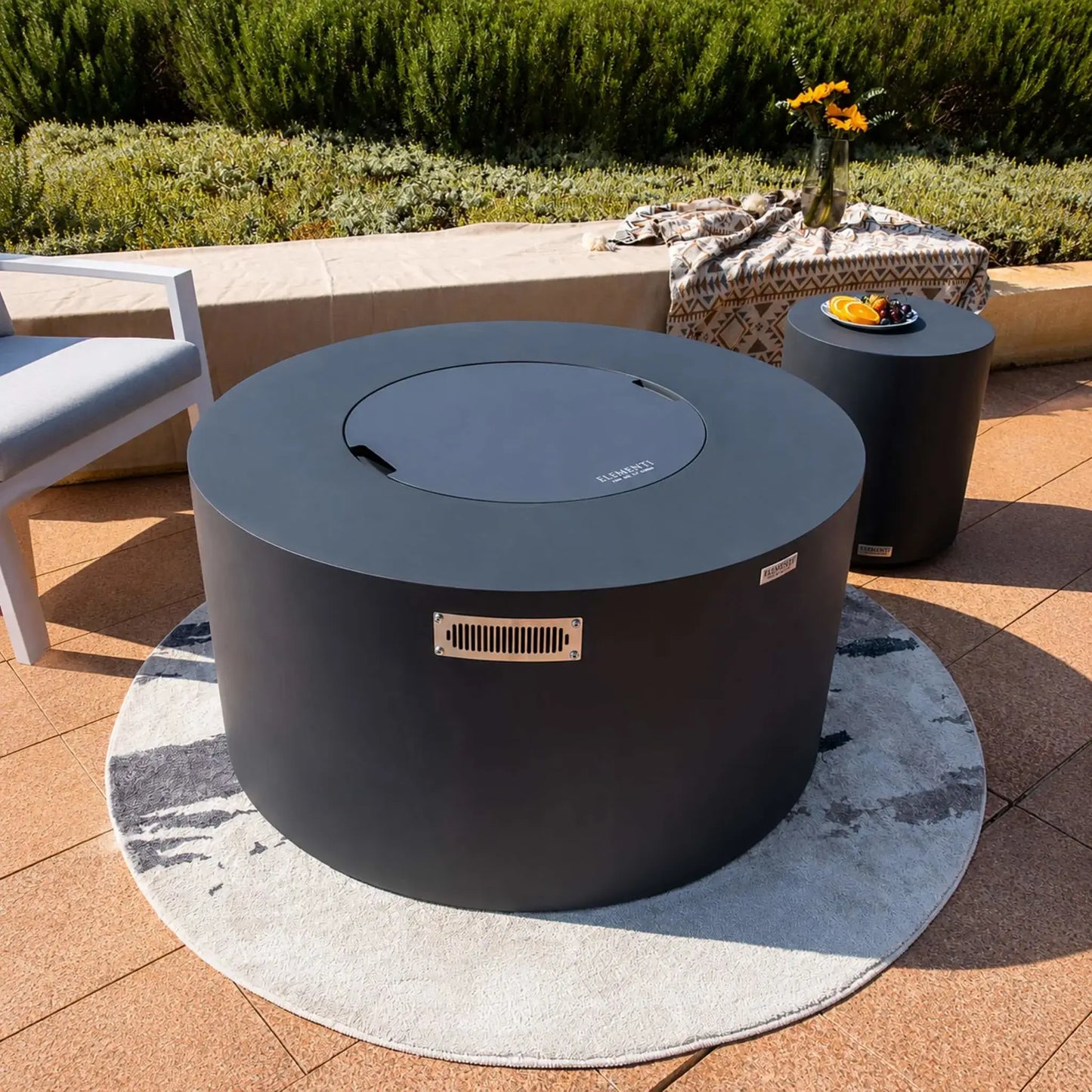 Wide lifestyle view of the Elementi Art of Living Pacaya Dark Grey round GFRC concrete fire pit table with the lid installed, transforming the fire feature into a functional outdoor table. The hidden propane fire pit sits on a circular area rug surrounded by modern patio seating, a matching cylindrical side table, decorative textiles, and lush waterfront landscaping, highlighting its contemporary outdoor living design.