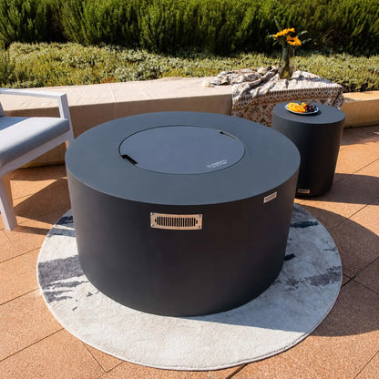 Wide lifestyle view of the Elementi Art of Living Pacaya Dark Grey round GFRC concrete fire pit table with the lid installed, transforming the fire feature into a functional outdoor table. The hidden propane fire pit sits on a circular area rug surrounded by modern patio seating, a matching cylindrical side table, decorative textiles, and lush waterfront landscaping, highlighting its contemporary outdoor living design.