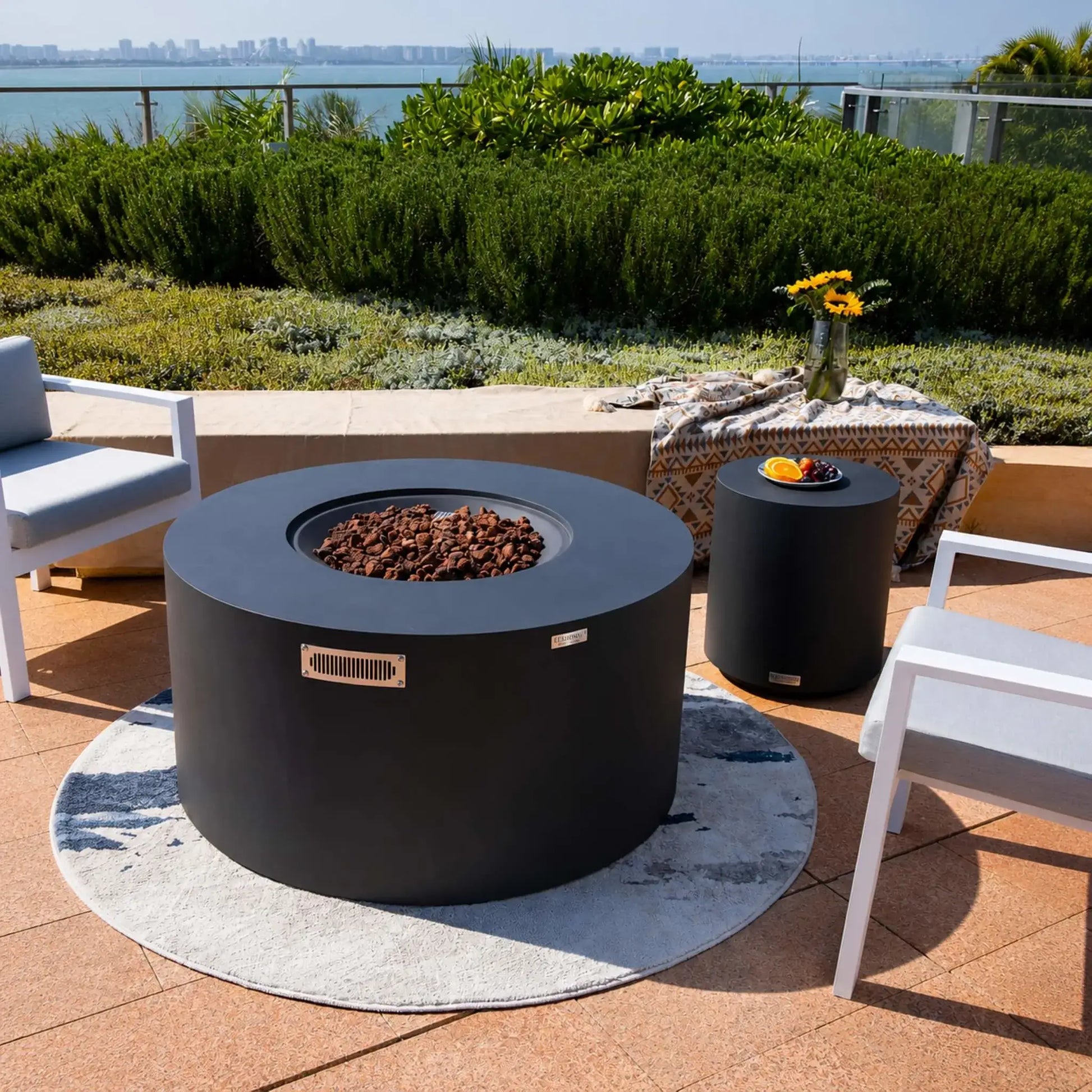 Elementi Art of Living Pacaya Dark Grey round GFRC concrete fire pit table with the burner cover removed, revealing a recessed lava rock fire bowl. Set on a waterfront patio surrounded by modern lounge chairs and manicured landscaping, the hidden propane fire pit blends luxury outdoor heating with contemporary furniture styling for upscale residential entertaining spaces.