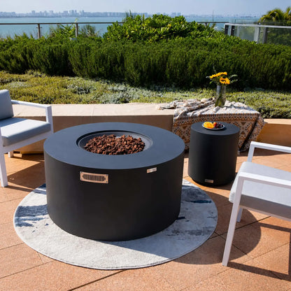Elementi Art of Living Pacaya Dark Grey round GFRC concrete fire pit table with the burner cover removed, revealing a recessed lava rock fire bowl. Set on a waterfront patio surrounded by modern lounge chairs and manicured landscaping, the hidden propane fire pit blends luxury outdoor heating with contemporary furniture styling for upscale residential entertaining spaces.