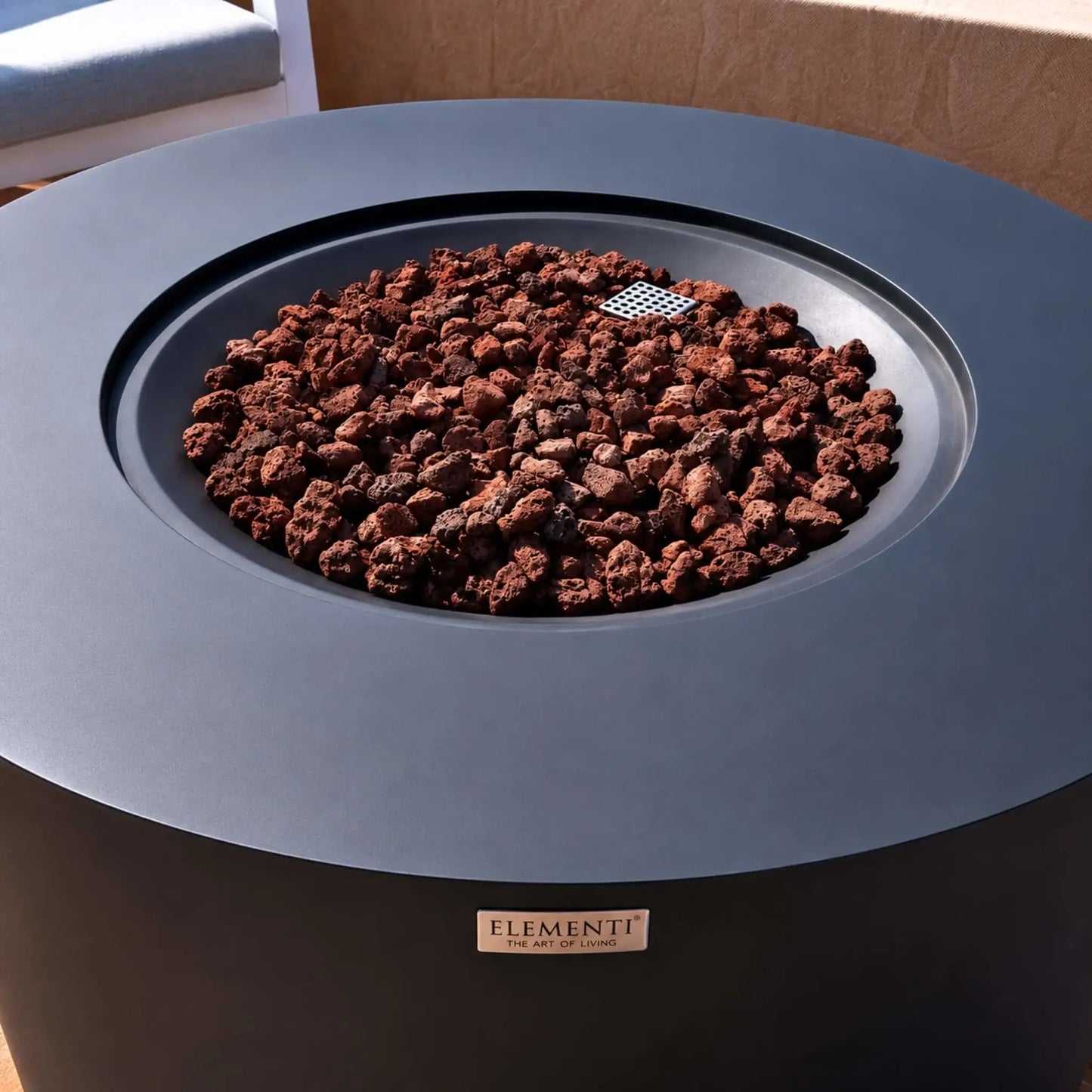 Detailed close-up of the Elementi Art of Living Pacaya Dark Grey round fire pit table fire bowl filled with natural lava rock. The recessed burner area is framed by a smooth GFRC concrete tabletop, highlighting the premium construction, hidden propane fire pit design, and contemporary outdoor aesthetic suitable for luxury patios, decks, and entertainment spaces.
