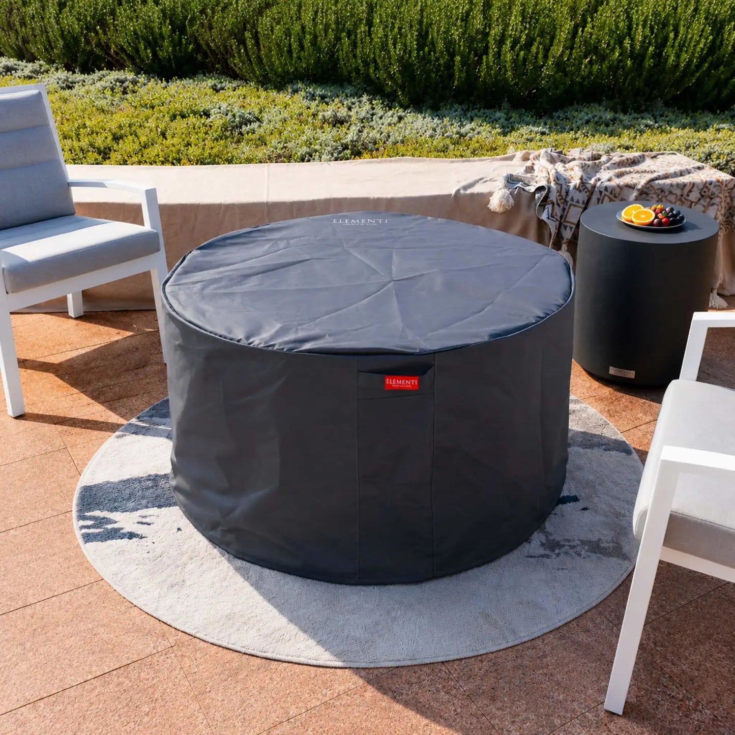 Elementi Art of Living Pacaya Dark Grey round fire pit table protected with a fitted weather-resistant cover. The cover encloses the GFRC concrete fire pit table while maintaining its cylindrical profile, demonstrating off-season protection and long-term outdoor durability. The covered hidden propane fire pit remains integrated within a stylish patio lounge setting overlooking landscaped gardens.