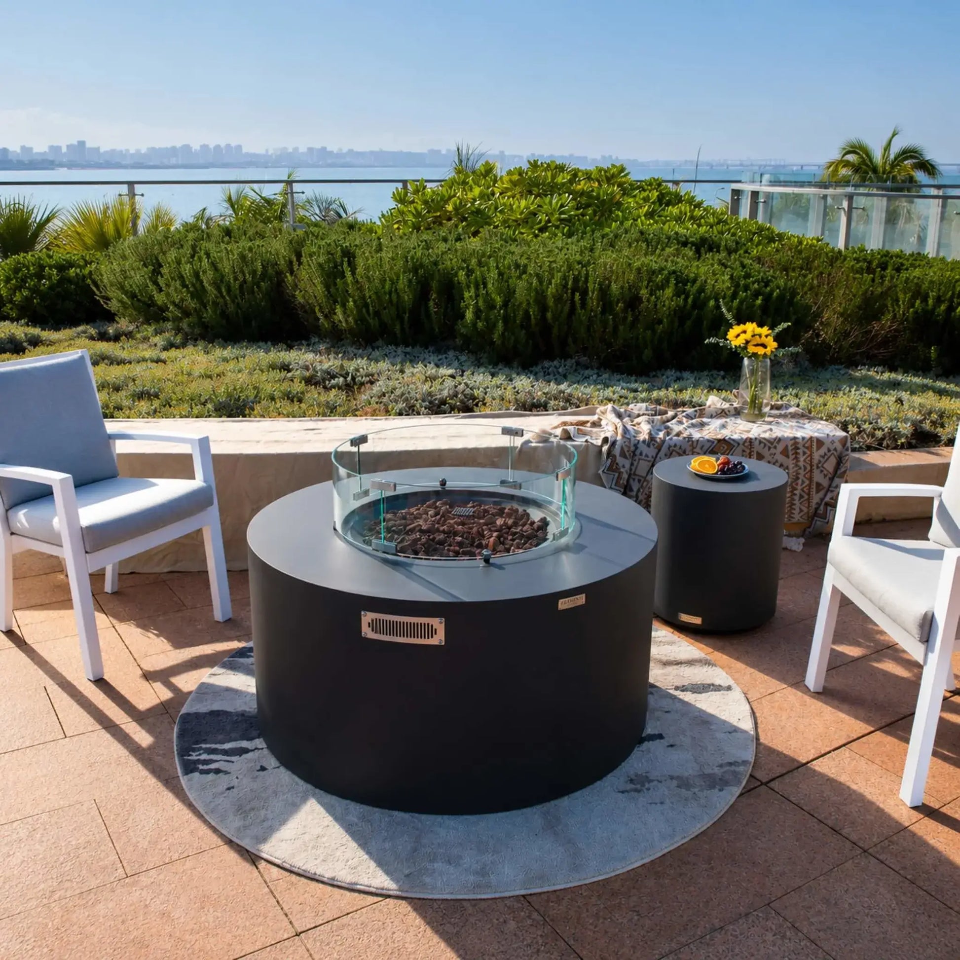 Elementi Art of Living Pacaya Dark Grey round fire pit table equipped with a clear tempered glass wind guard surrounding the lava rock burner area. Positioned on a luxury waterfront patio with modern seating and a matching side table, the hidden propane fire pit combines safety, flame protection, and contemporary outdoor design for premium entertaining environments.
