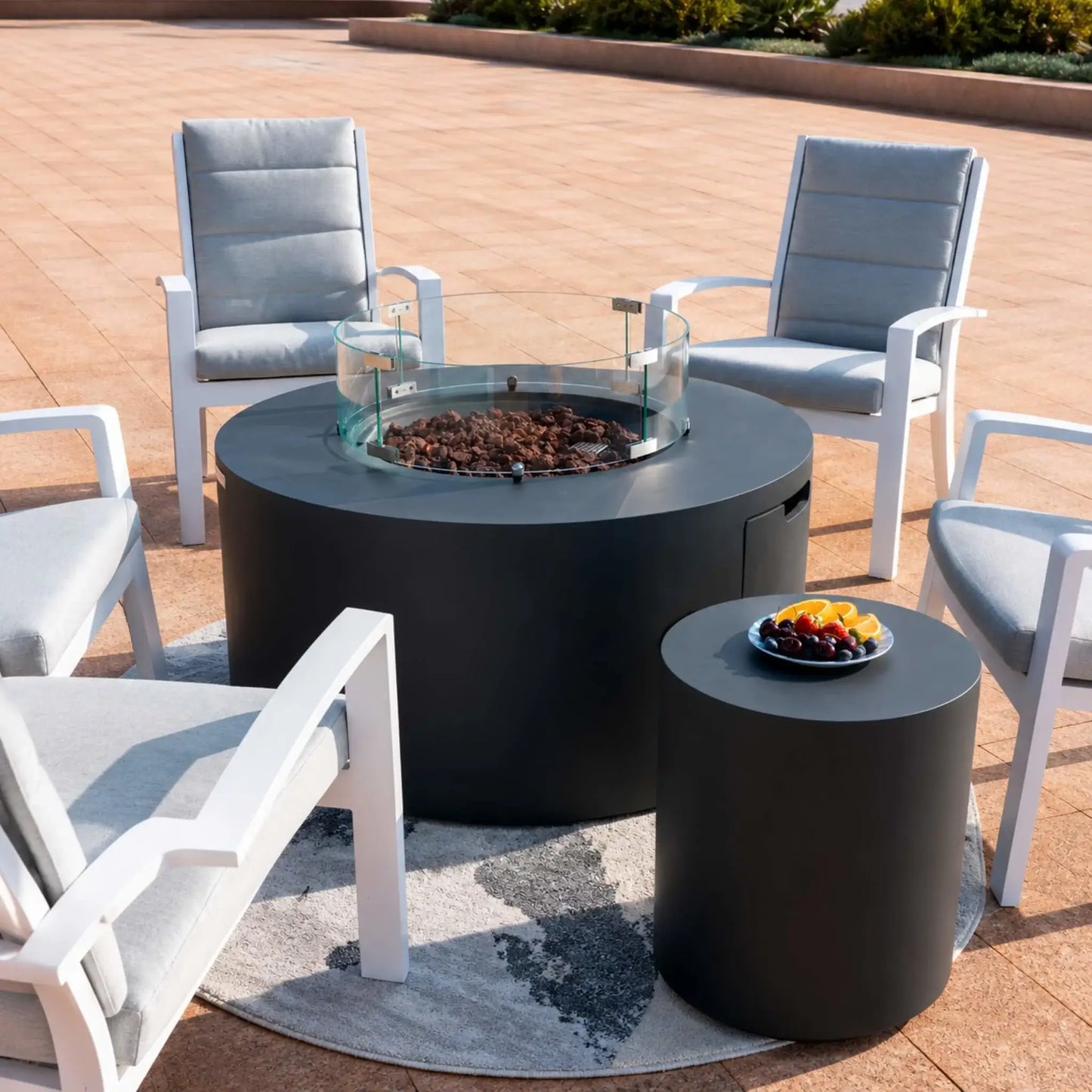 Angled patio view of the Elementi Art of Living Pacaya Dark Grey round GFRC concrete fire pit table featuring a tempered glass wind guard and recessed lava rock fire bowl. Surrounded by cushioned outdoor armchairs and a coordinating side table, the hidden propane fire pit creates an inviting conversation area while showcasing modern circular furniture styling.