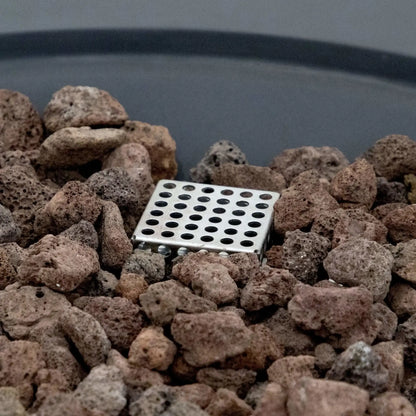 Macro close-up of the lava rock burner media inside the Elementi Art of Living Pacaya Dark Grey fire pit table. The image focuses on the hidden propane burner component surrounded by natural volcanic rocks within the recessed fire bowl, highlighting the materials and functional design of this round GFRC concrete outdoor fire pit table.