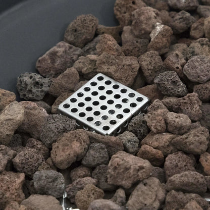 Close-up view of the stainless steel burner cover within the lava rock media of the Elementi Art of Living Pacaya Dark Grey round GFRC concrete fire pit table. The image highlights the hidden propane burner component surrounded by natural volcanic lava rocks, showcasing the premium construction and functional fire media design of this modern outdoor fire pit table.